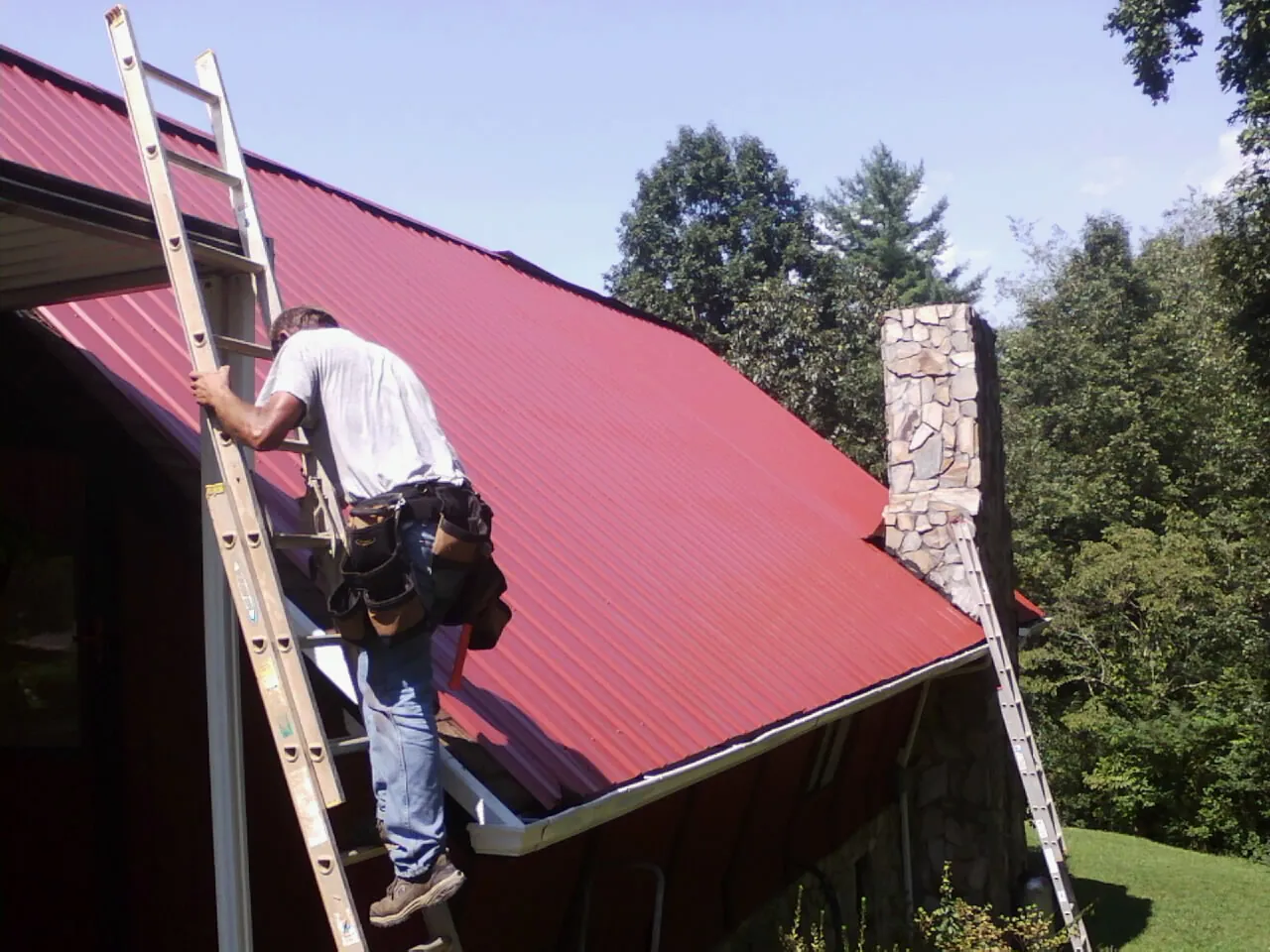 Roofer on a ladder beside a red metal roof and stone chimney — Soffit & Fascia Repair by Roofing Brothers in Otisville